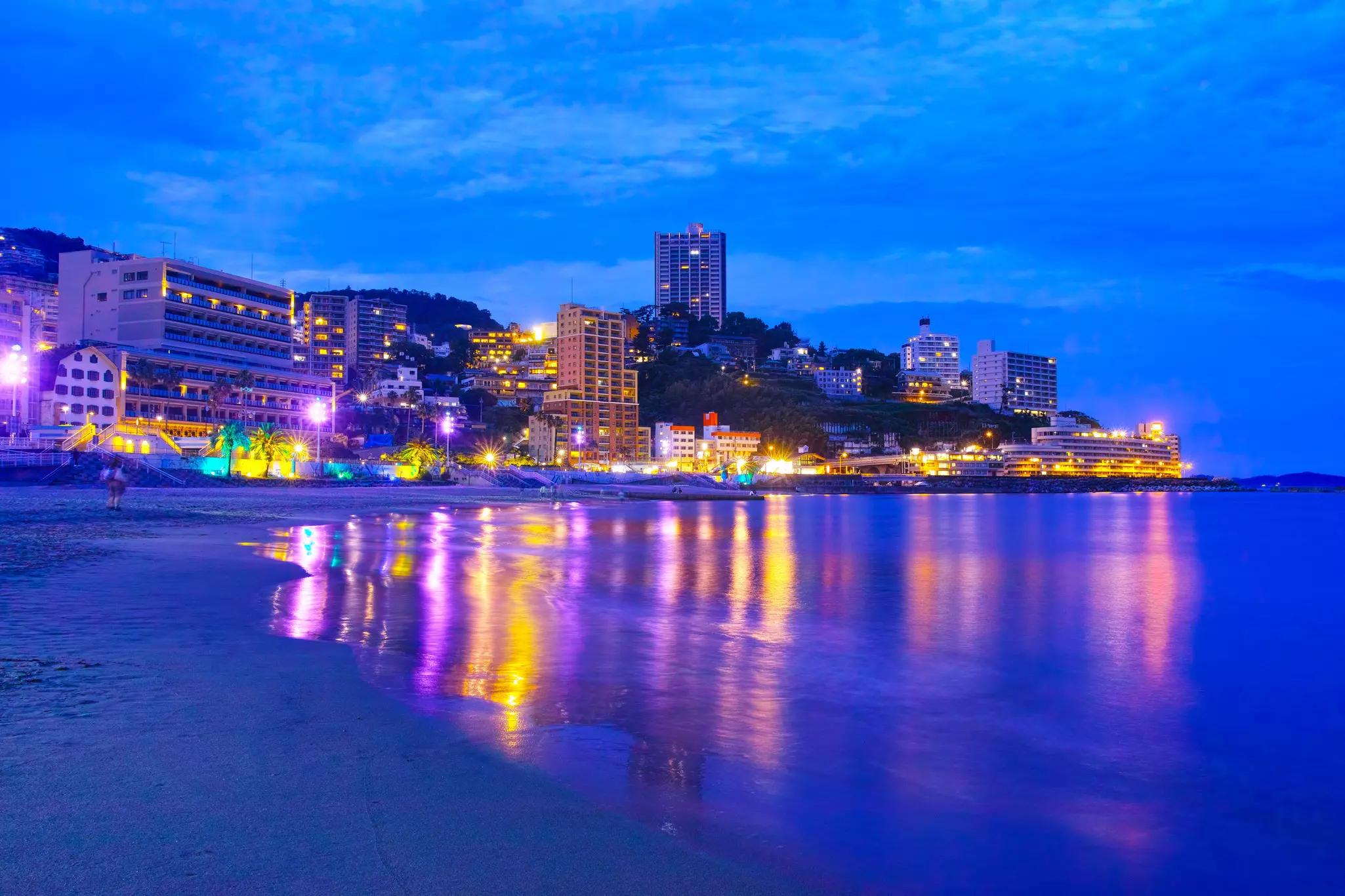 Buildings with lights are reflected in the water at night on a beach.