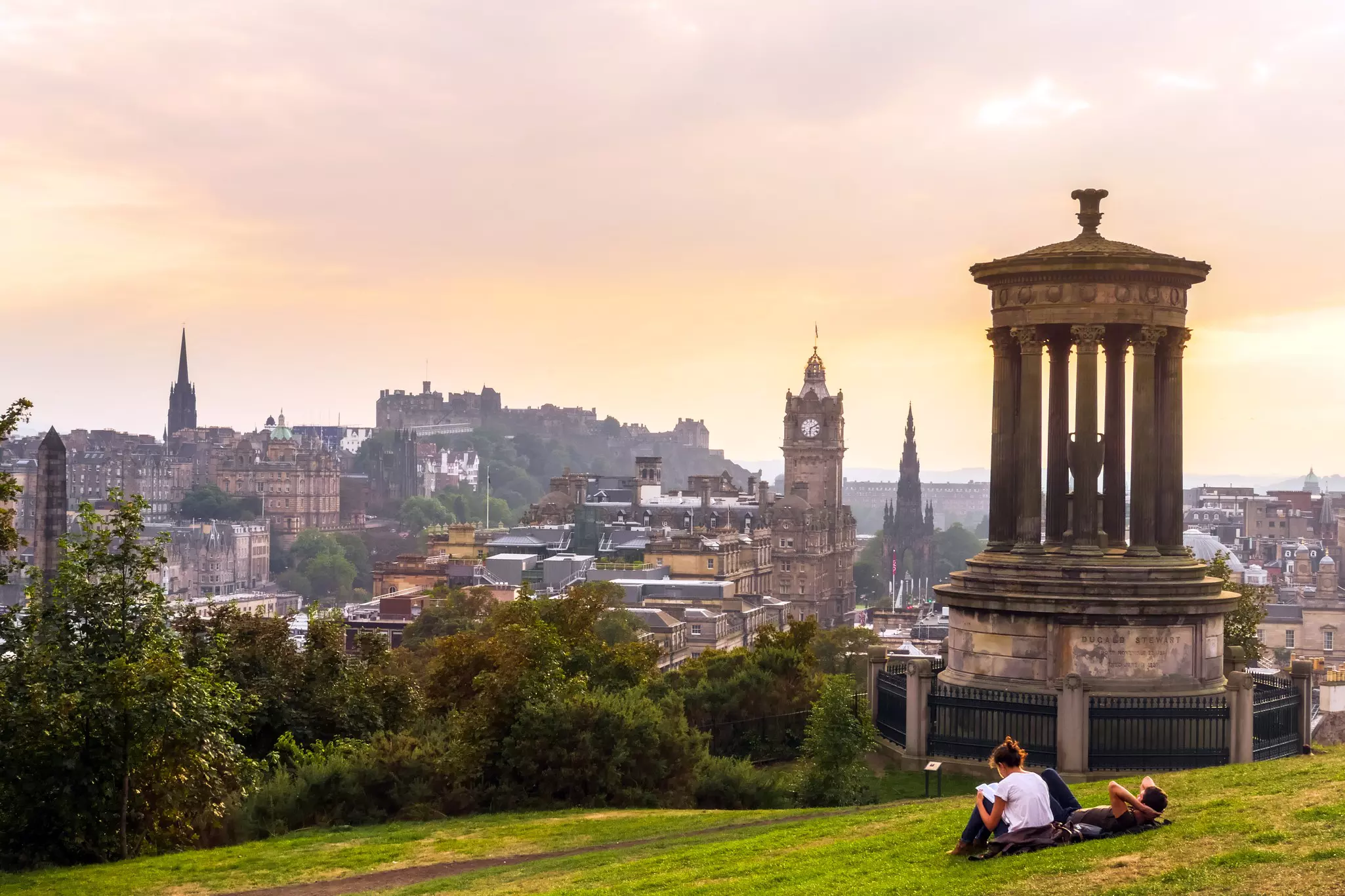 A couple sit on a hill with a city view below including a clock tower, church steeples and a hilltop castle