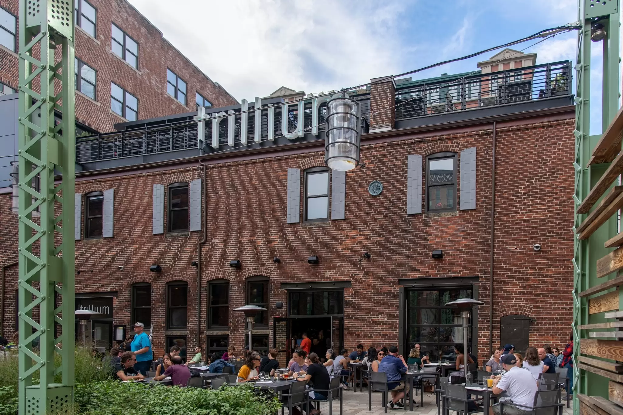 People eating and drinking at outdoor tables in the courtyard of a large, redbrick brewery.