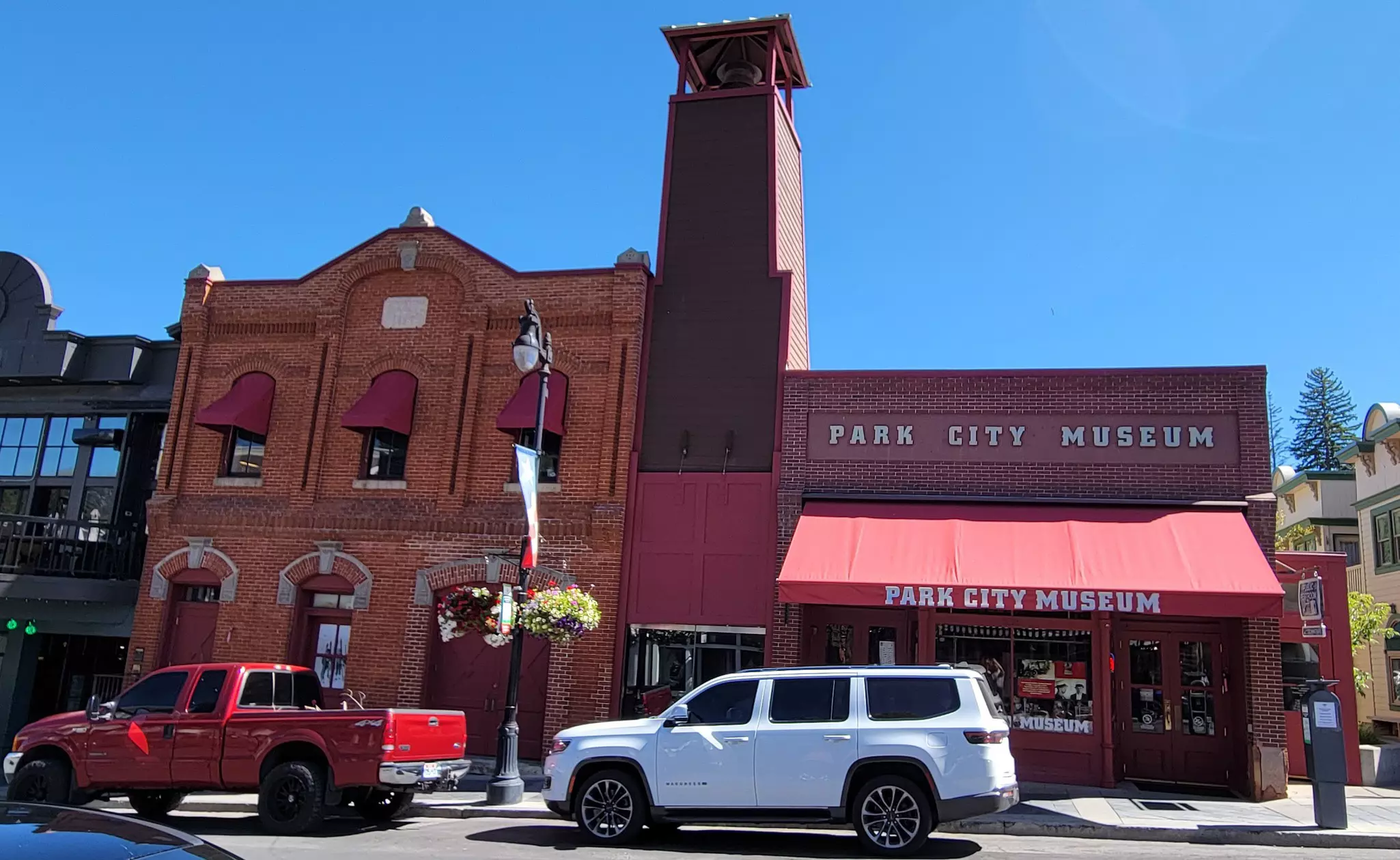 Exterior of low brick buildings on a city street. The building to the right has an awning and lettering above, both reading "Park City Museum." An SUV and pickup truck are parked out front on a sunny day.