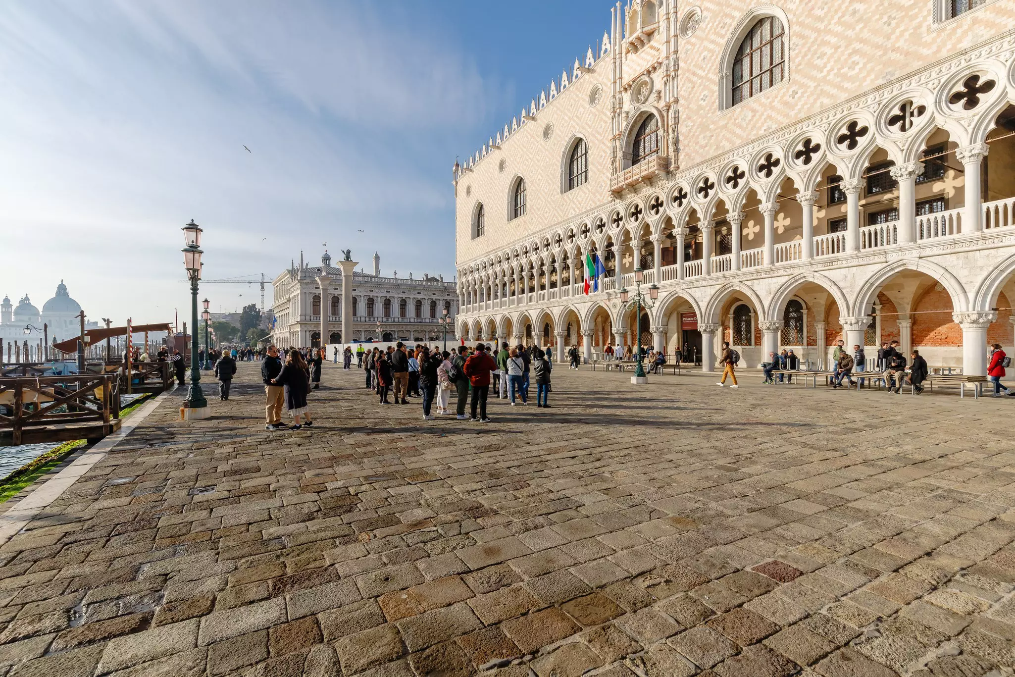 Groups of people outside a grand building with arches in its facade at the edge of a canal.