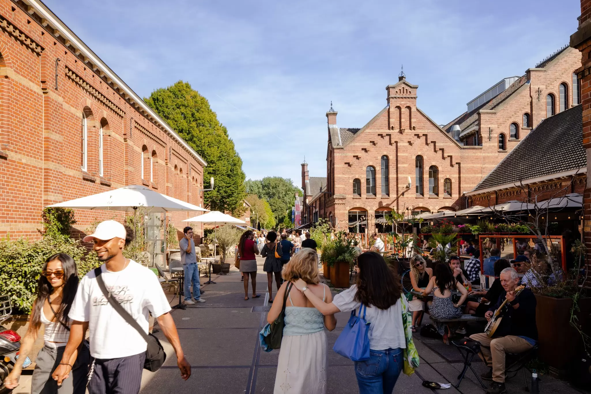 People walk past the brick buildings of the Cultural Park Westergas in Amsterdam, Netherlands, on a sunny day. 