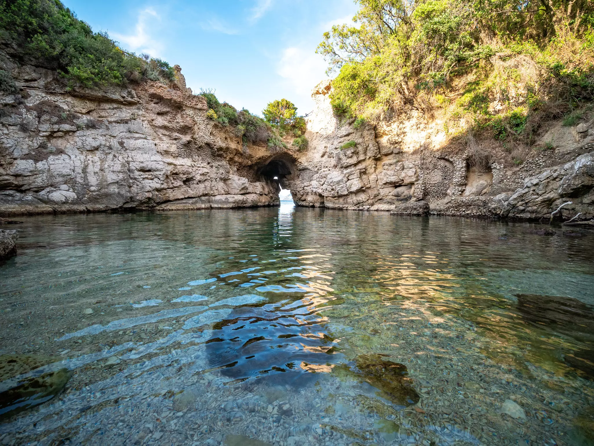A shallow swimming hole enclosed by high rocks with an arched opening to the sea beyond on a sunny day.