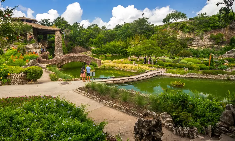 A couple stands at a bridge over a pond in a lushly green Japanese-style garden in San Antonio, Texas.