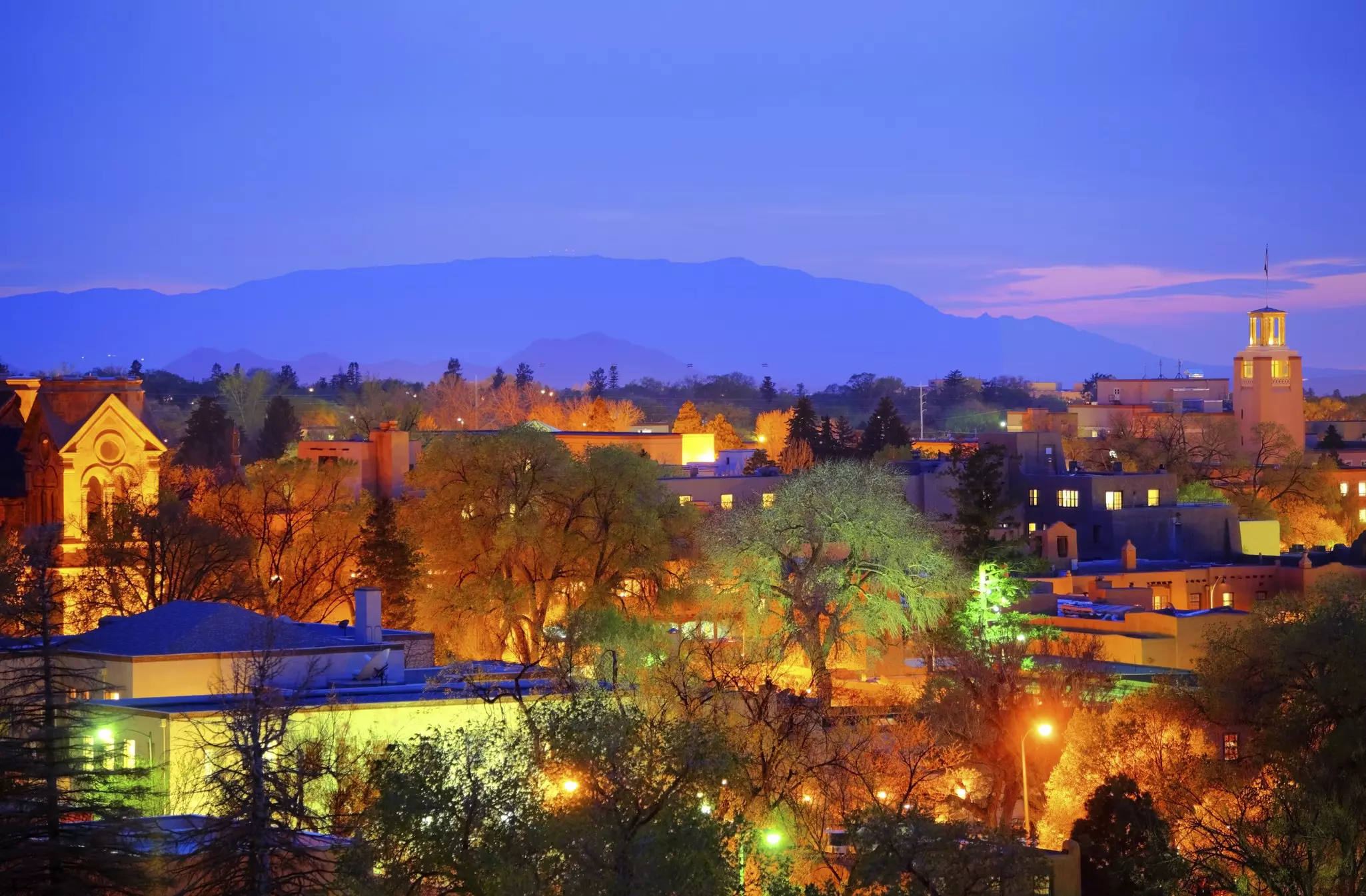 The illuminated low-rise skyline of downtown Santa Fe at dusk, with pink streaks of light against the mountain range in the background