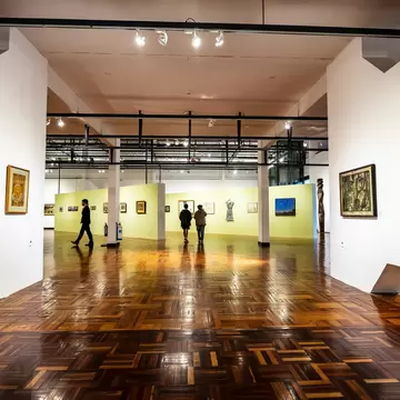 Visitors walk through a white-walled gallery hung with framed paintings above a polished parquet floor.