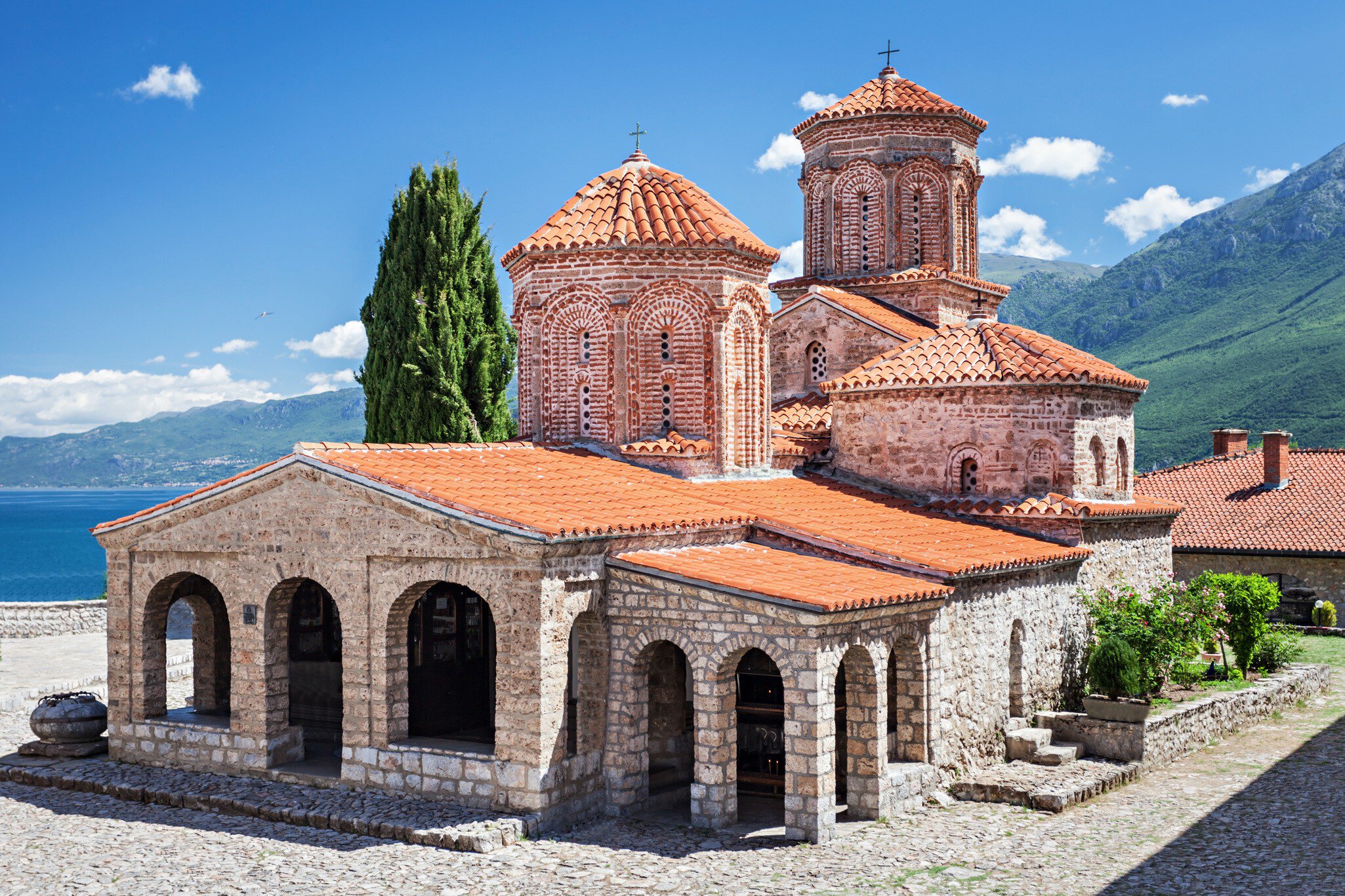 Stone church with red-tiled domes and an arched porch beside a tall cypress, blue lake and green mountains behind