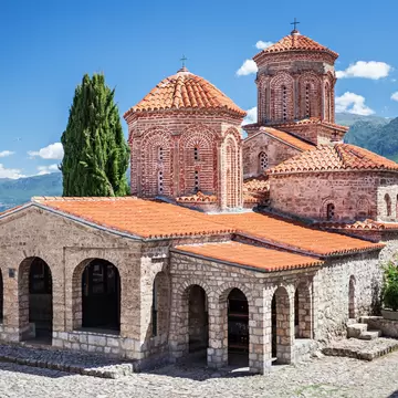 Stone church with red-tiled domes and an arched porch beside a tall cypress, blue lake and green mountains behind