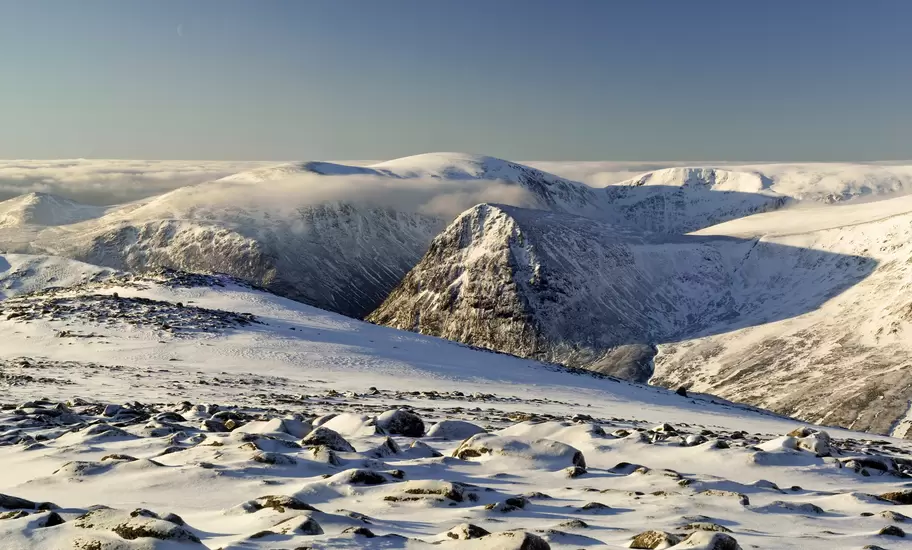 Snow-covered rounded mountains and a rocky, boulder-strewn plateau under a clear blue winter sky, with low cloud in the valleys.