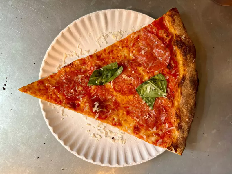Top-down close-up of a triangular slice of pizza with spicy salami and basil leaf on the metal counter at L'Industrie pizzeria on Christopher Street in the West Village, New York City