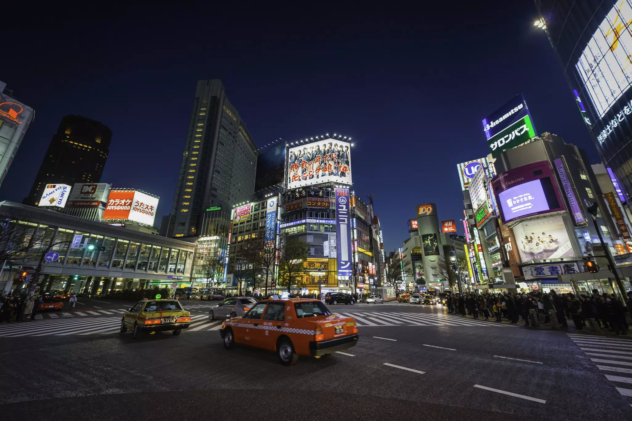 Red taxis are seen in an intersection of a city surrounded by billboards and lights.