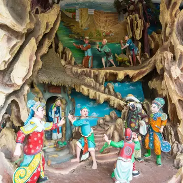Brightly costumed statues arranged in a grotto-like tableau with a painted river scene behind, at Haw Par Villa in Singapore.