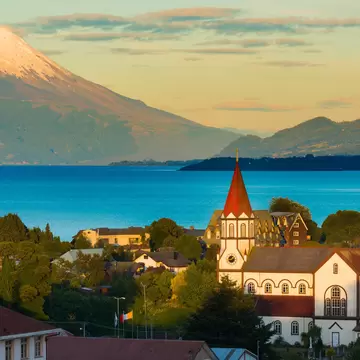 White church with red spires among trees in Puerto Varas, above a blue lake and a snow-capped volcano lit by low sun.
