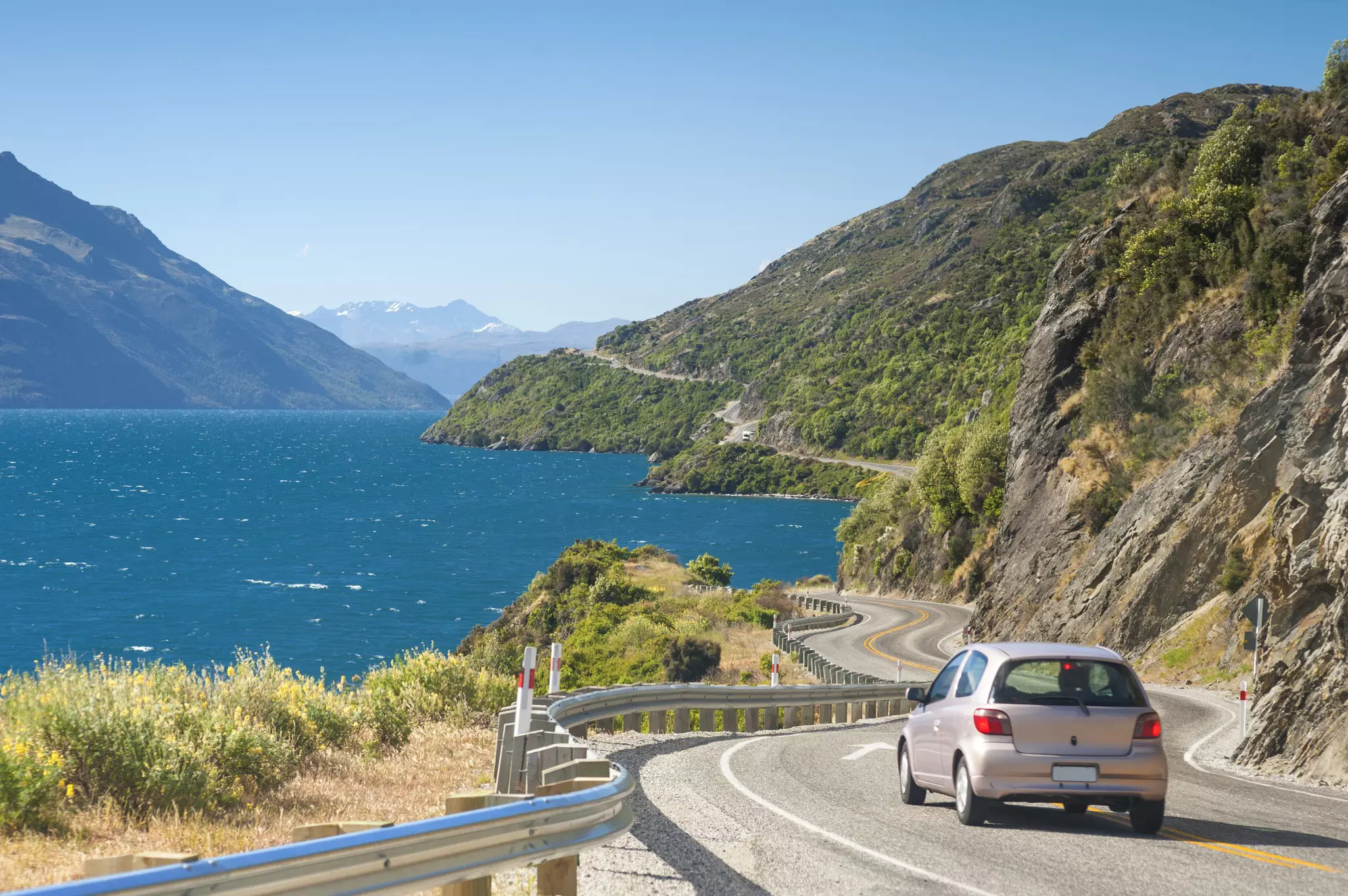 A small gray car drives on a two-lane road by a lake in New Zealand; hills rise from the shoreline.