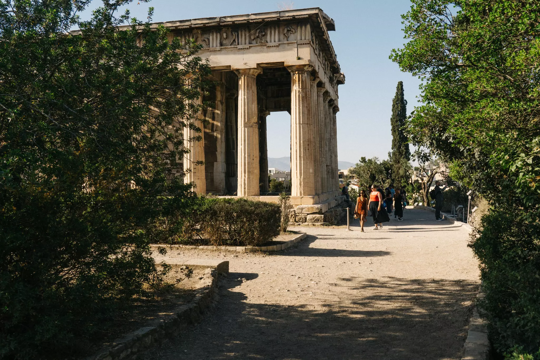 Tourists walk on a path lined with greenery past an ancient white stone building with columns.