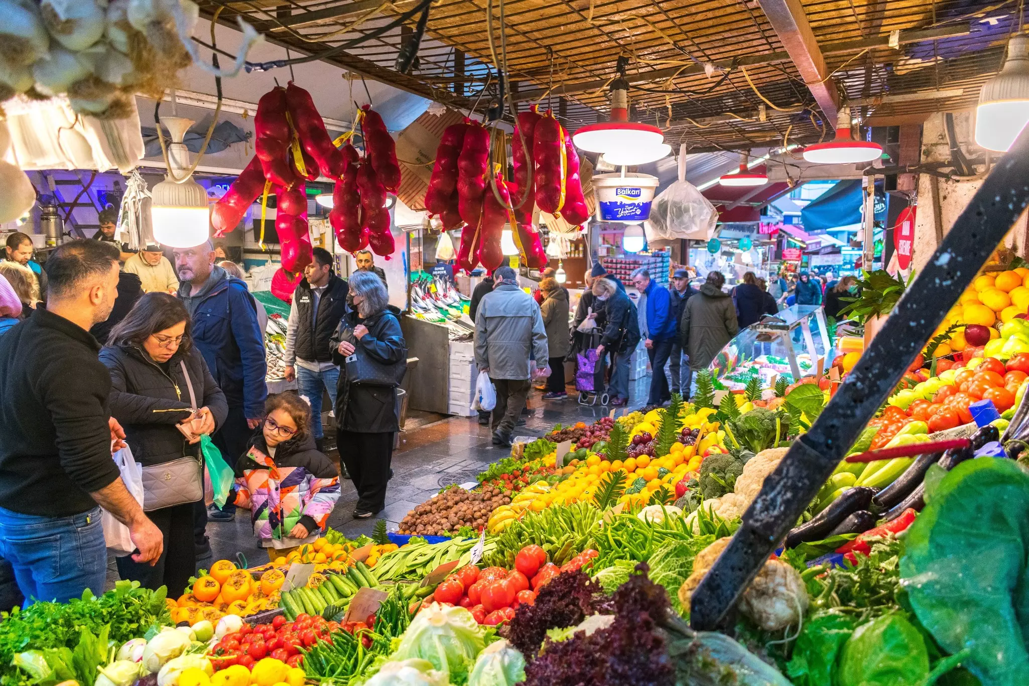 People shop in a fresh food market at a stall full of colorful fruits and vegetables.