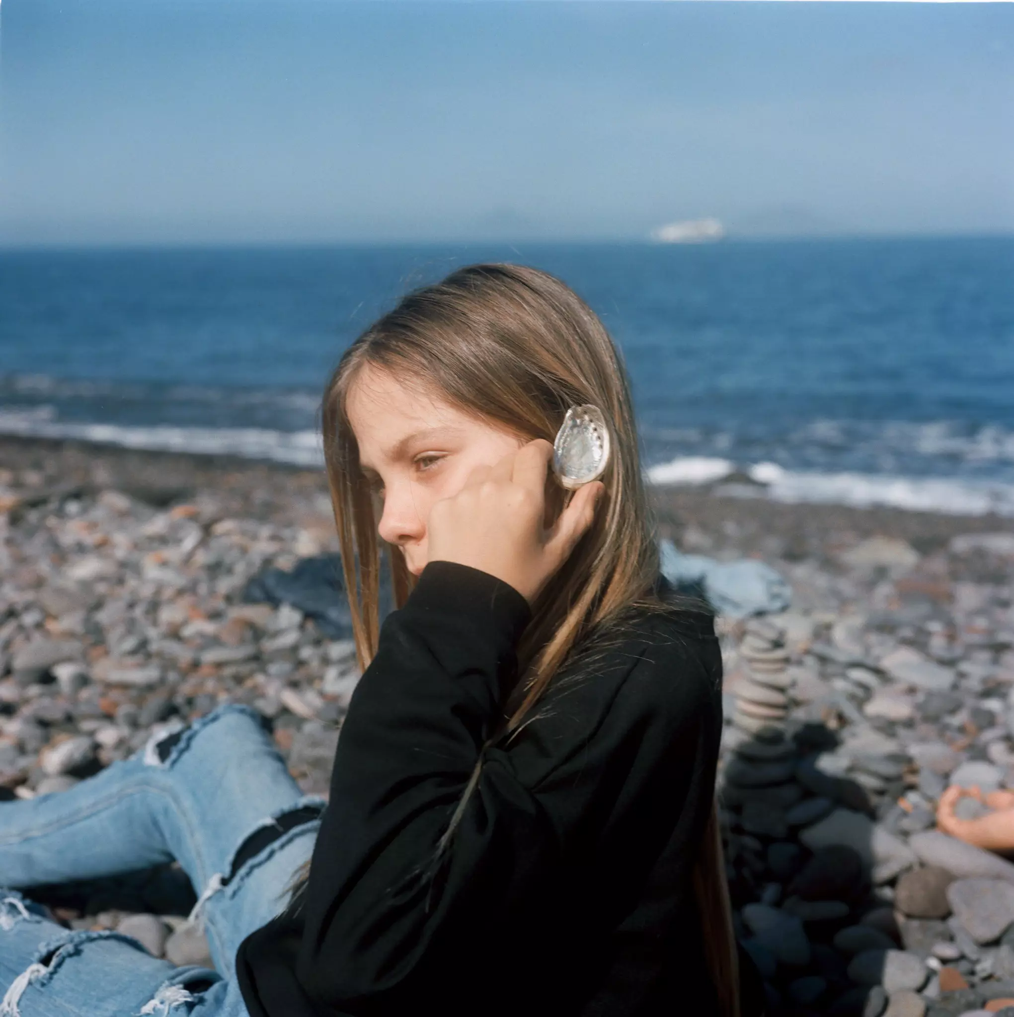 A girl sitting on a rocky beach holds a clear plastic to her ear.
