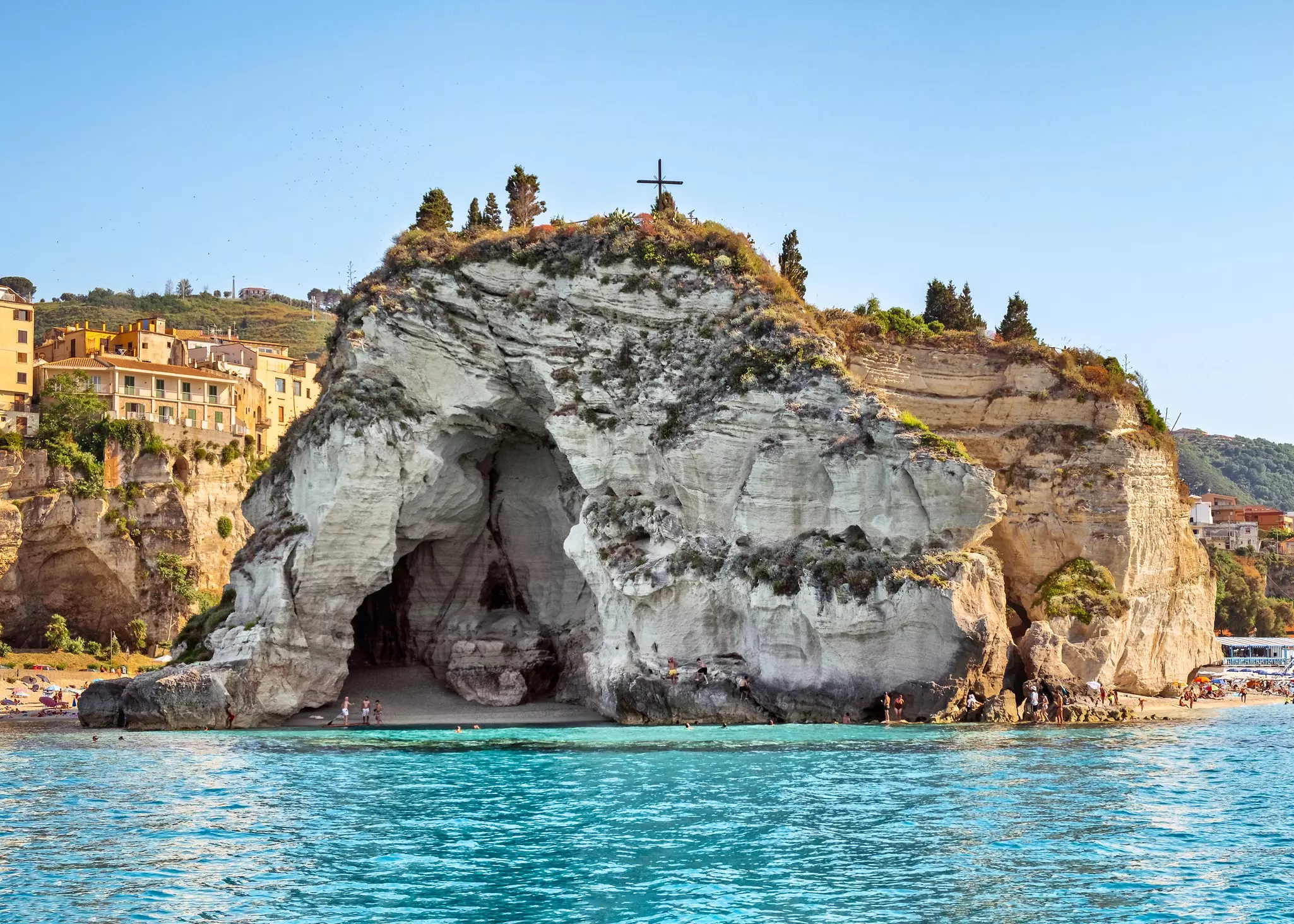 A cave within a cliff with people heading towards the sandy beach.
