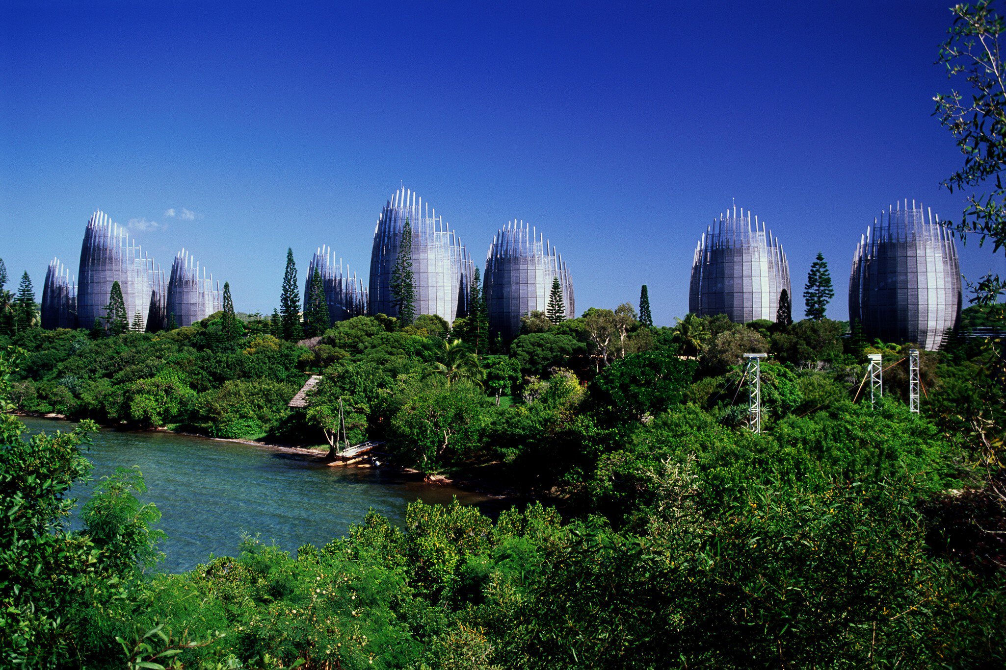A row of tall curved timber-and-steel pavilions shaped like woven baskets, rising from dense green vegetation above a lagoon.