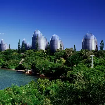 A row of tall curved timber-and-steel pavilions shaped like woven baskets, rising from dense green vegetation above a lagoon.