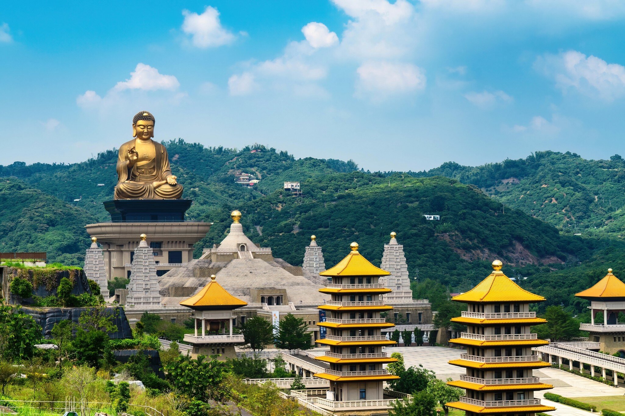 Golden buddha at the end of a row of yellow and gray pavilion buildings surrounded by green hillsides in Kaohsiung, Taiwan.