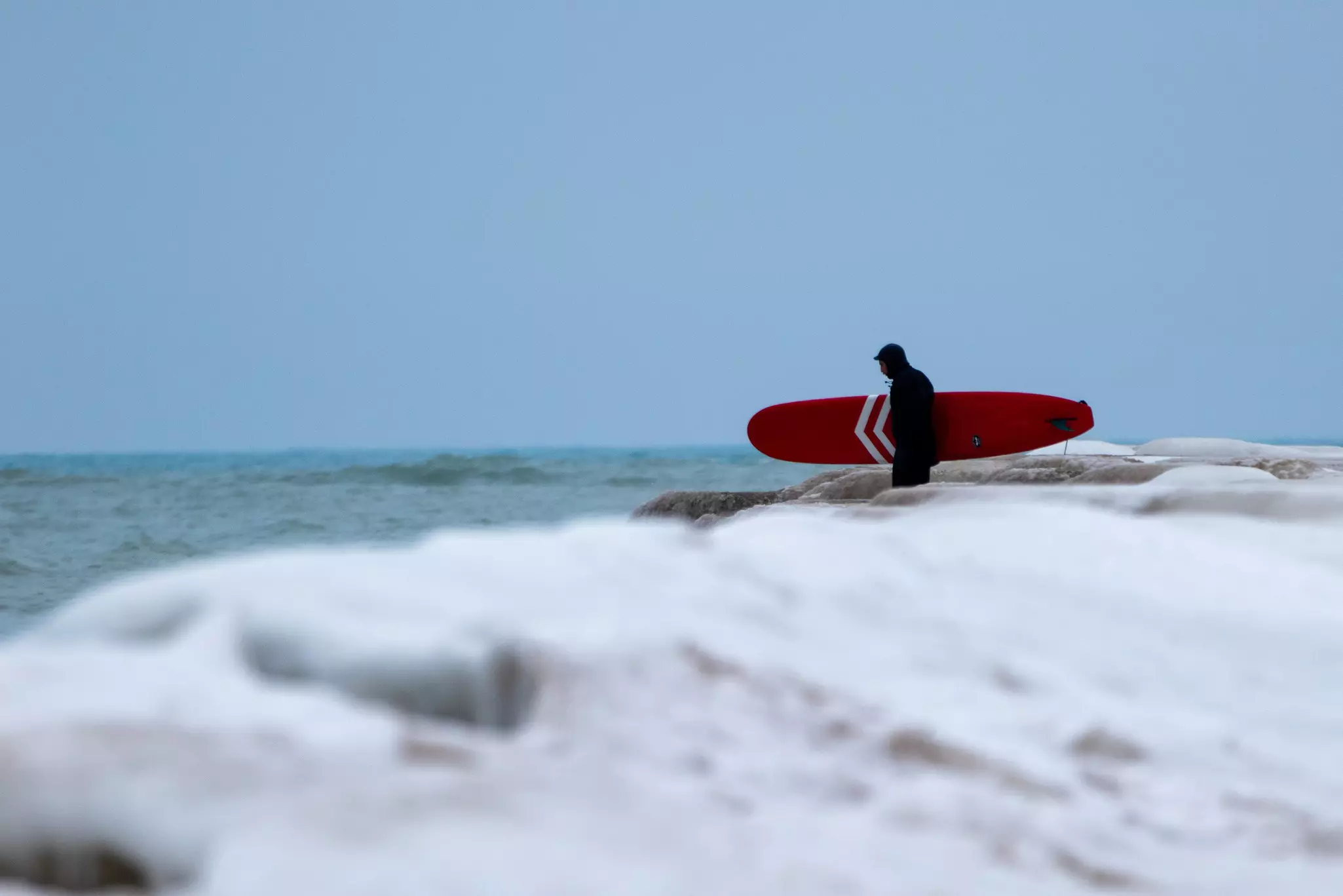 A person carries a red surfboard toward a lake with out-of-focus snow in the foreground on an overcast day.