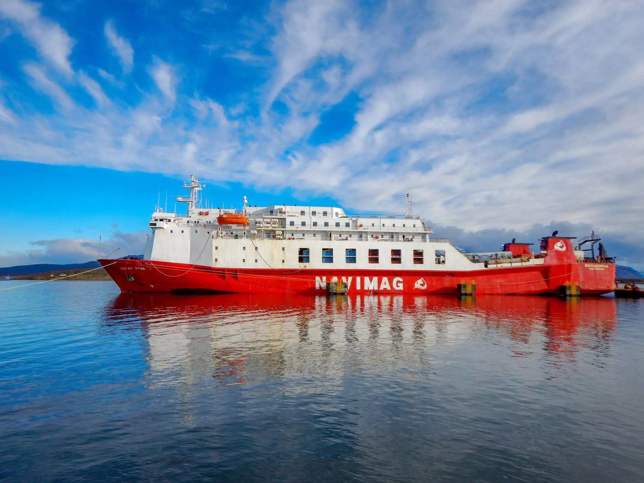 Large red and white ferry with "Navimag" in white letters on the side anchored in a harbor on a sunny day.