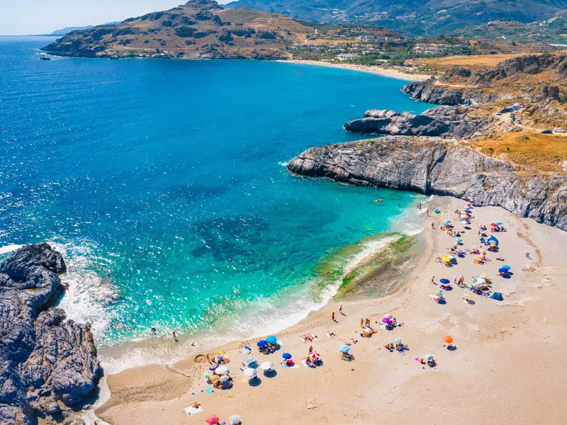 People with umbrellas relax on a sandy beach with clear blue water on Crete. 
