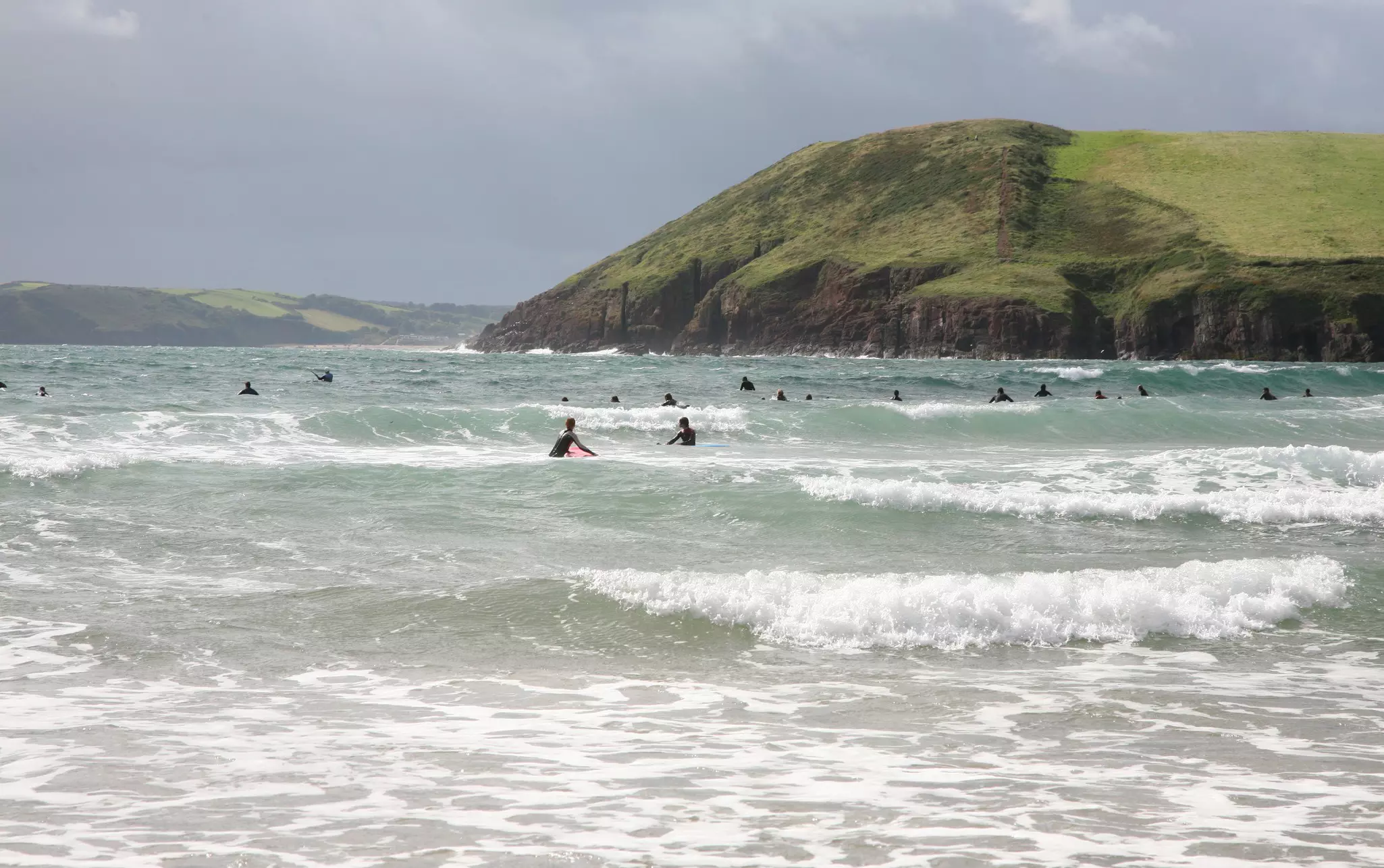 Surfers waiting for waves in sea with landscape and grey sky in background at Manorbier beach in Wales.