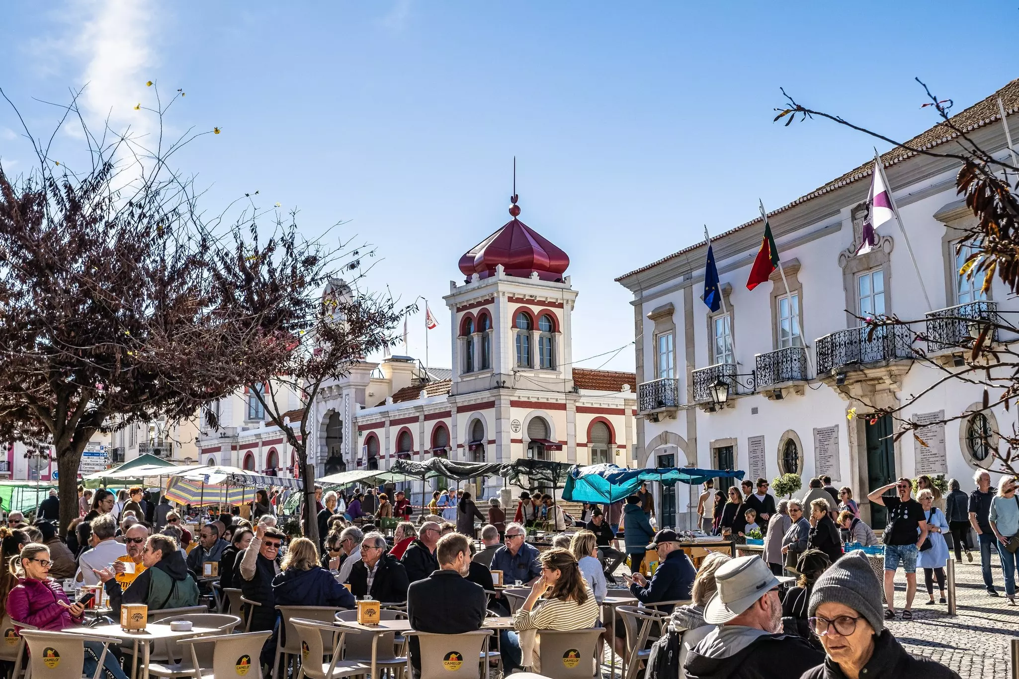 People sit at tables in a city square with a Moorish-style tower behind them.
