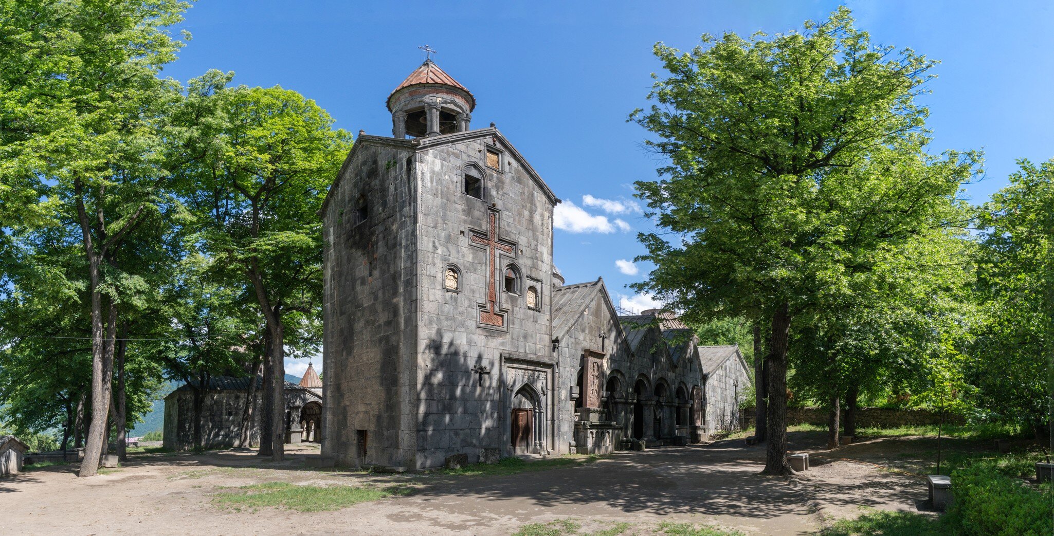 A weathered stone church with a carved cross and conical bell tower amid trees.