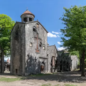 A weathered stone church with a carved cross and conical bell tower amid trees.