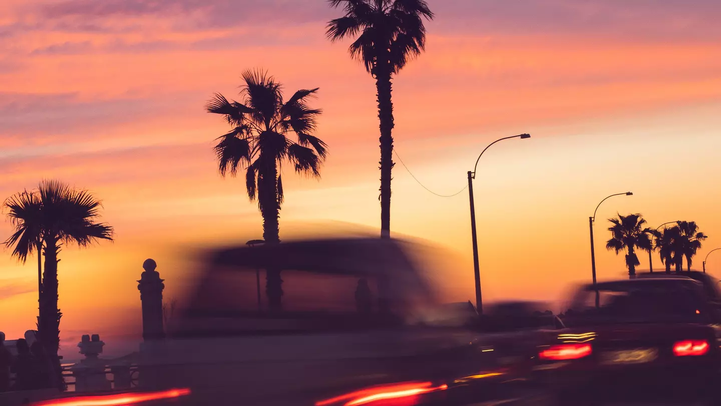 A long exposure of cars driving past palm trees, all silhouetted against a pink sunset sky