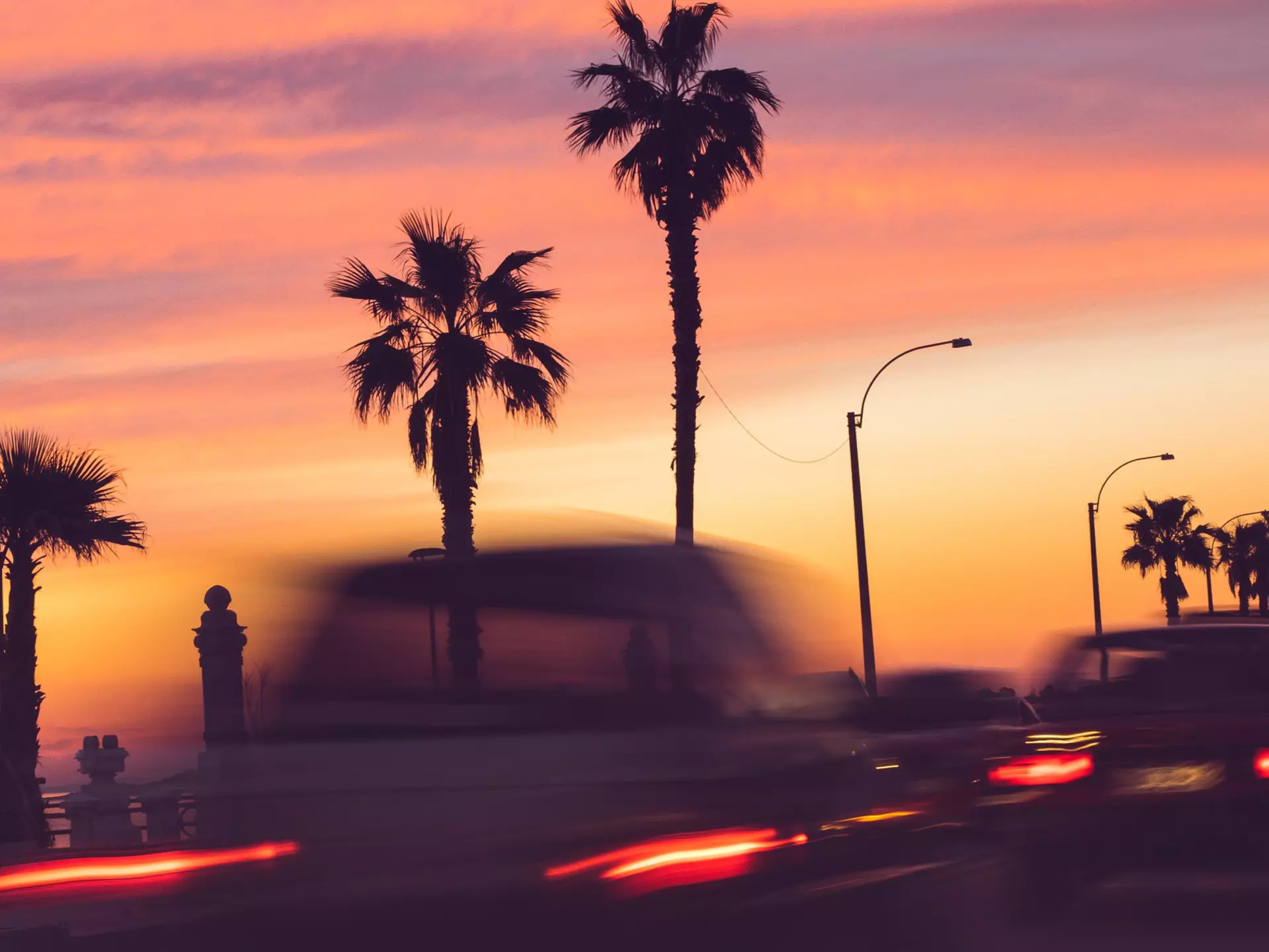 A long exposure of cars driving past palm trees, all silhouetted against a pink sunset sky