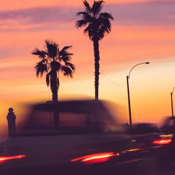 A long exposure of cars driving past palm trees, all silhouetted against a pink sunset sky