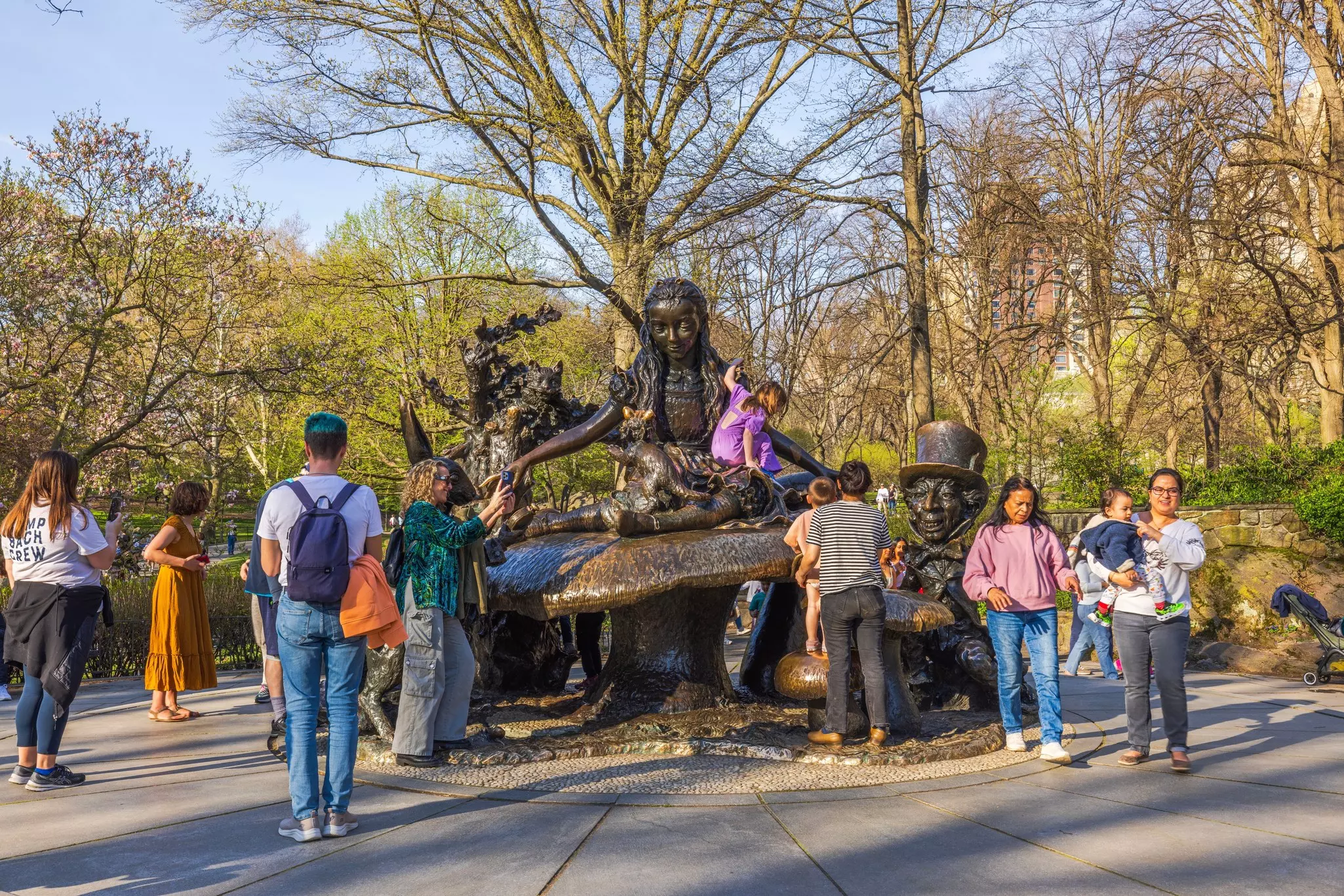 Parents watch as children climb on a huge bronze sculpture depicting a fairy-tale scene, set in an urban park.