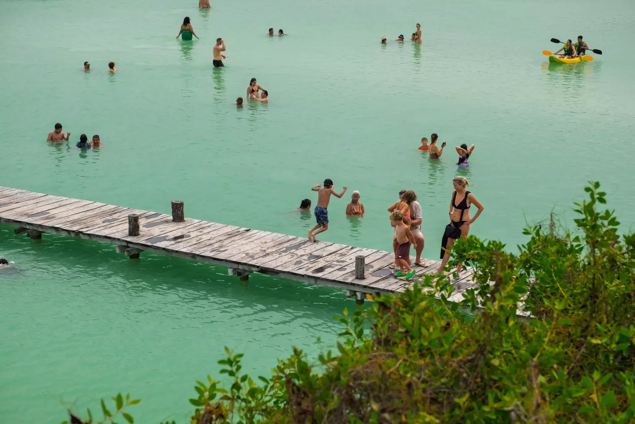 A person jumps from a long wooden pier into a greenish lake where people are standing in the water.