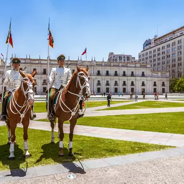 Two riders in white uniforms holding flagged lances sit on chestnut horses on a lawn before a long white facade