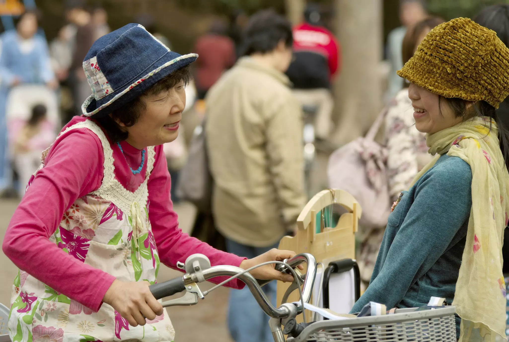 Two women, both wearing hats, talk on a street. The older of the two women sits on a bicycle.