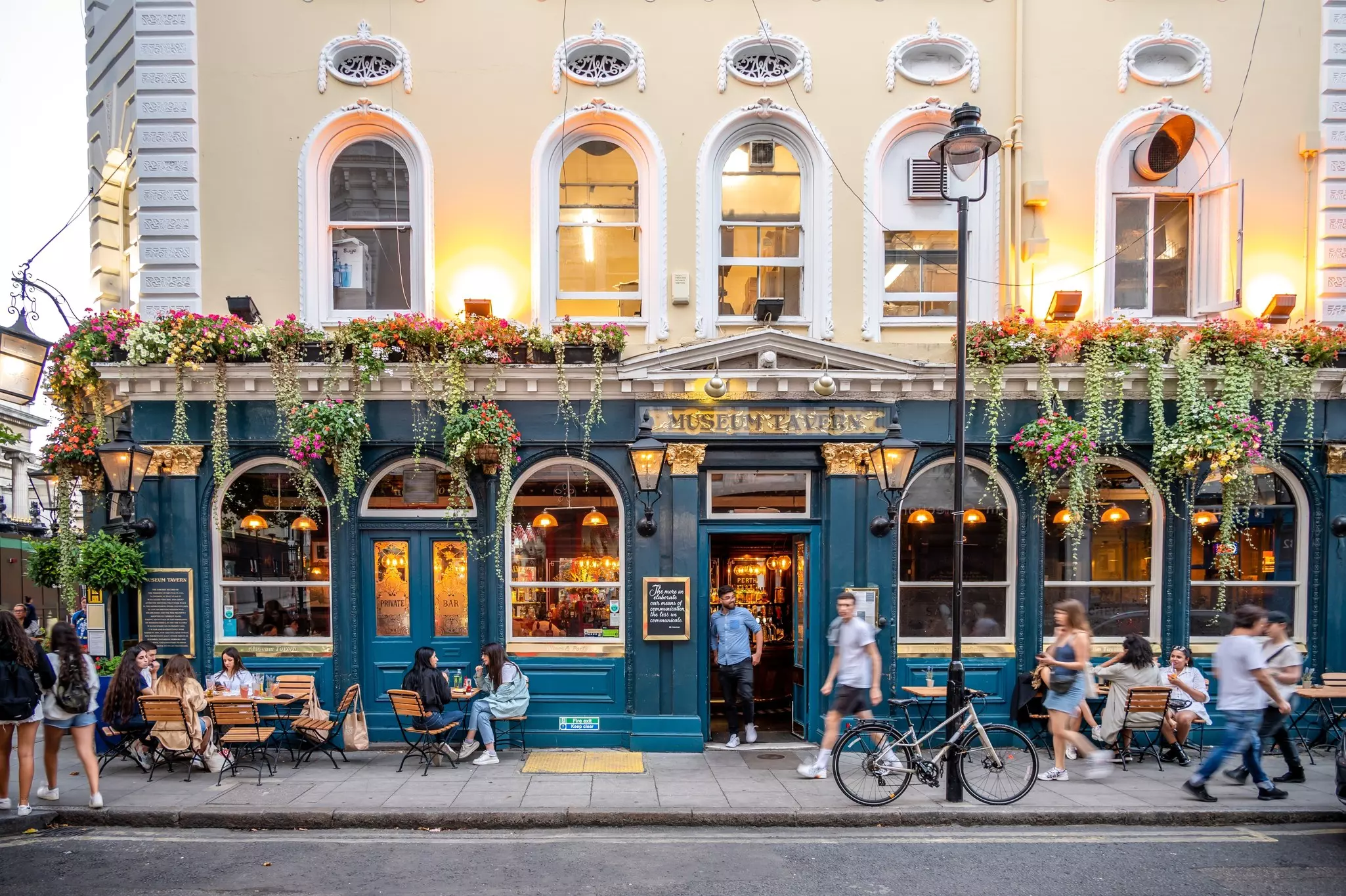 The exterior of a pub on a summer's evening, with people sat at tables on the pavement enjoying a drink. Baskets of flowers decorate the pub.
