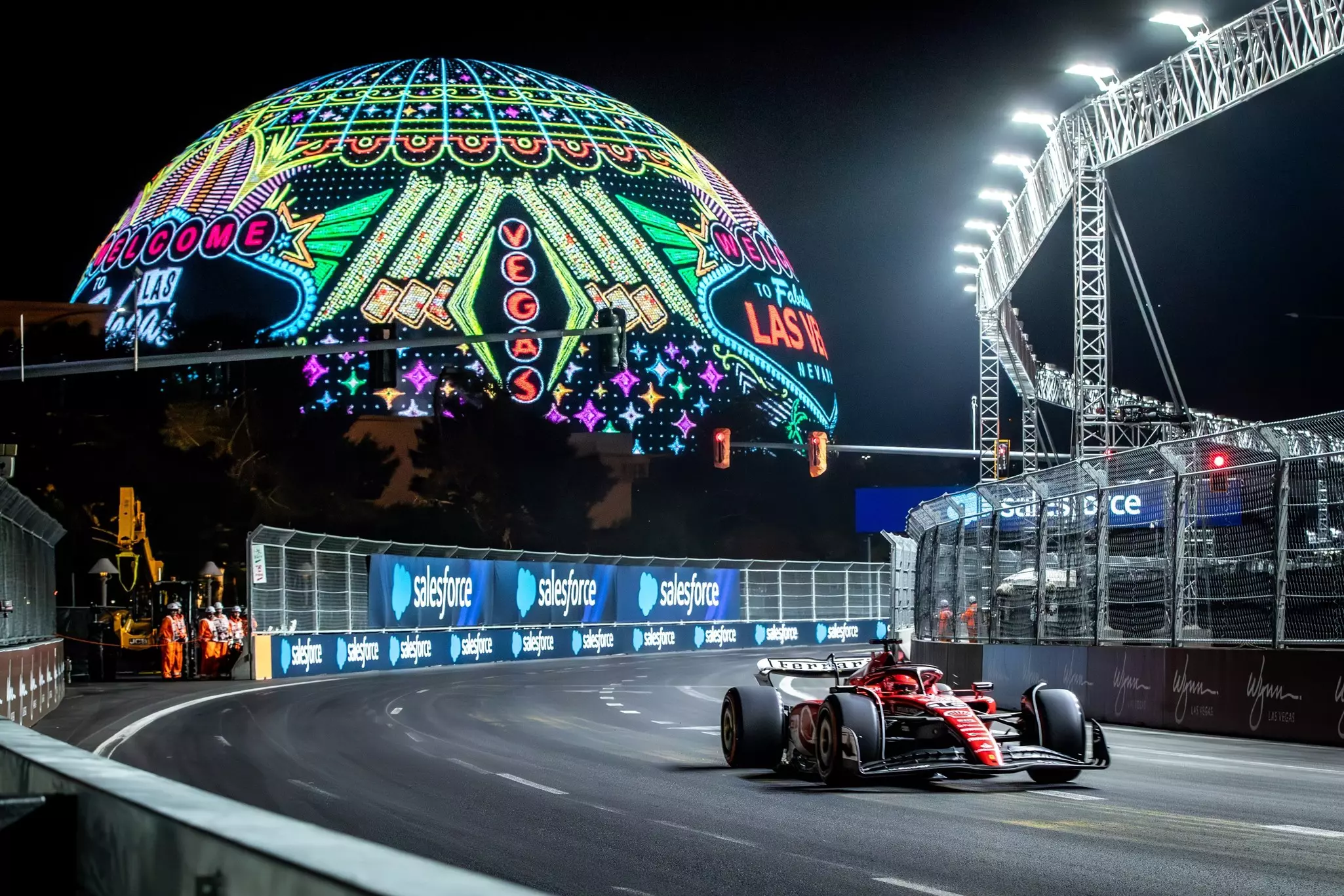 A race car speeds around a curve on a track at night. A huge, illuminated spherical structure is seen in the background.