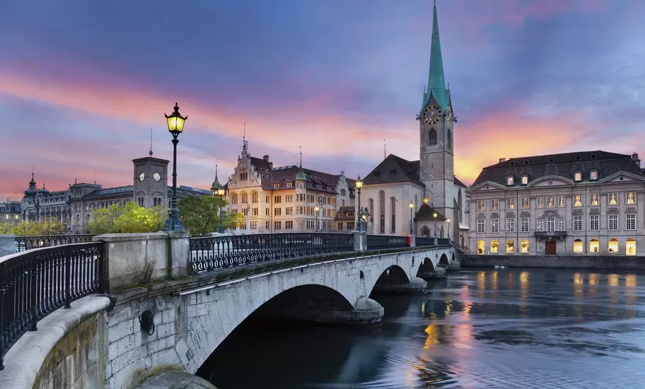 Zürich's Fraumünster spire and the arched Münsterbrücke over the Limmat River under a pink sunset sky, lamps lit