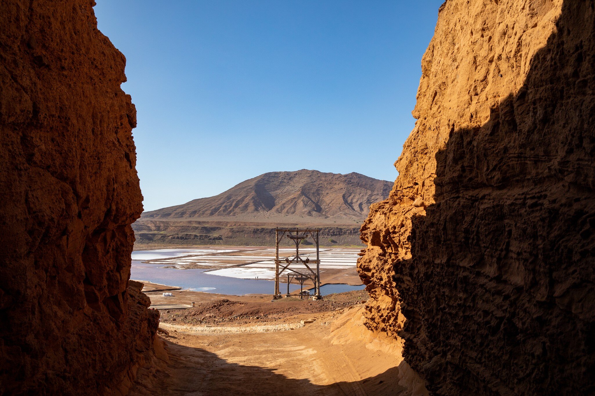 View through a sunlit rock passage to a wooden hoist frame, pale salt pans and a brown mountain beyond.