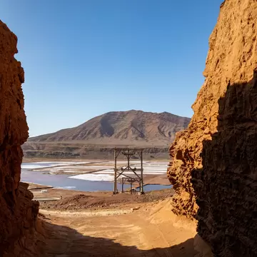 View through a sunlit rock passage to a wooden hoist frame, pale salt pans and a brown mountain beyond.