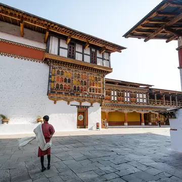 A local guide standing still while wearing a traditional clothing for men called Gho a knee length and kimono-like cloth held in place by a belt called Kera. Rinpung Dzong is an architectural feat housing a network of courtyards, temples and offic...