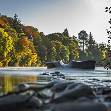 Carved wooden seat half submerged in a fast-flowing river, autumn woodland and a slate-roofed turret on the bank