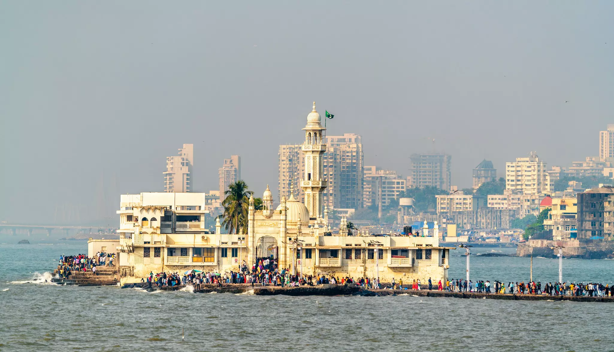 A white mosque on an island out at sea with many people walking across a causeway to visit.