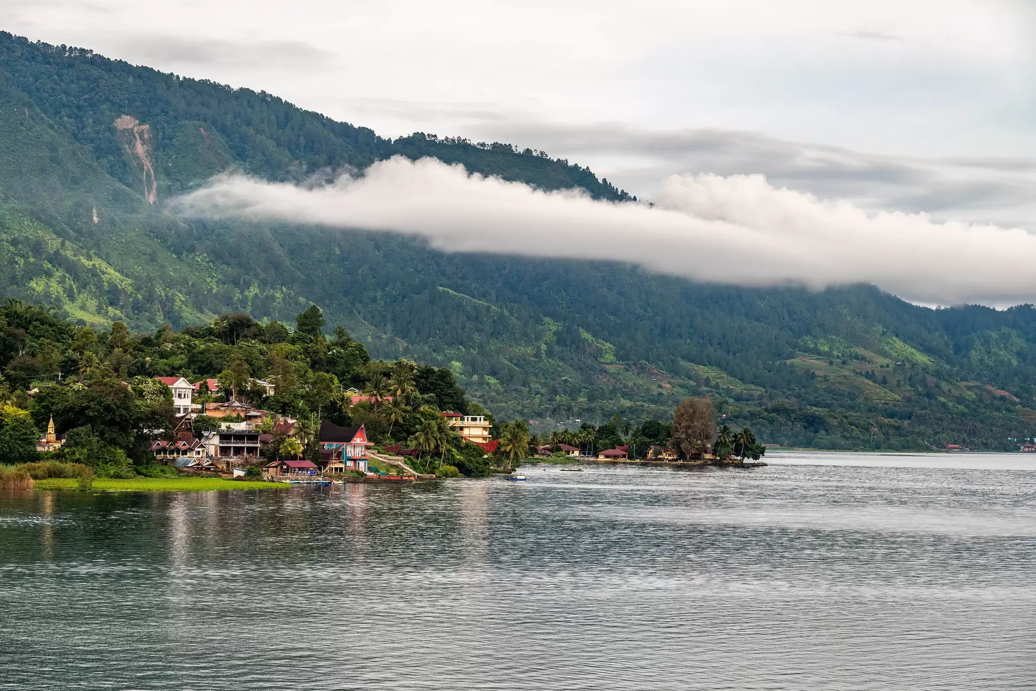 Low-hanging clouds are pictured over a shorefront village and steep green hillsides on Danau Toba (Lake Toba), Sumatra, Indonesia