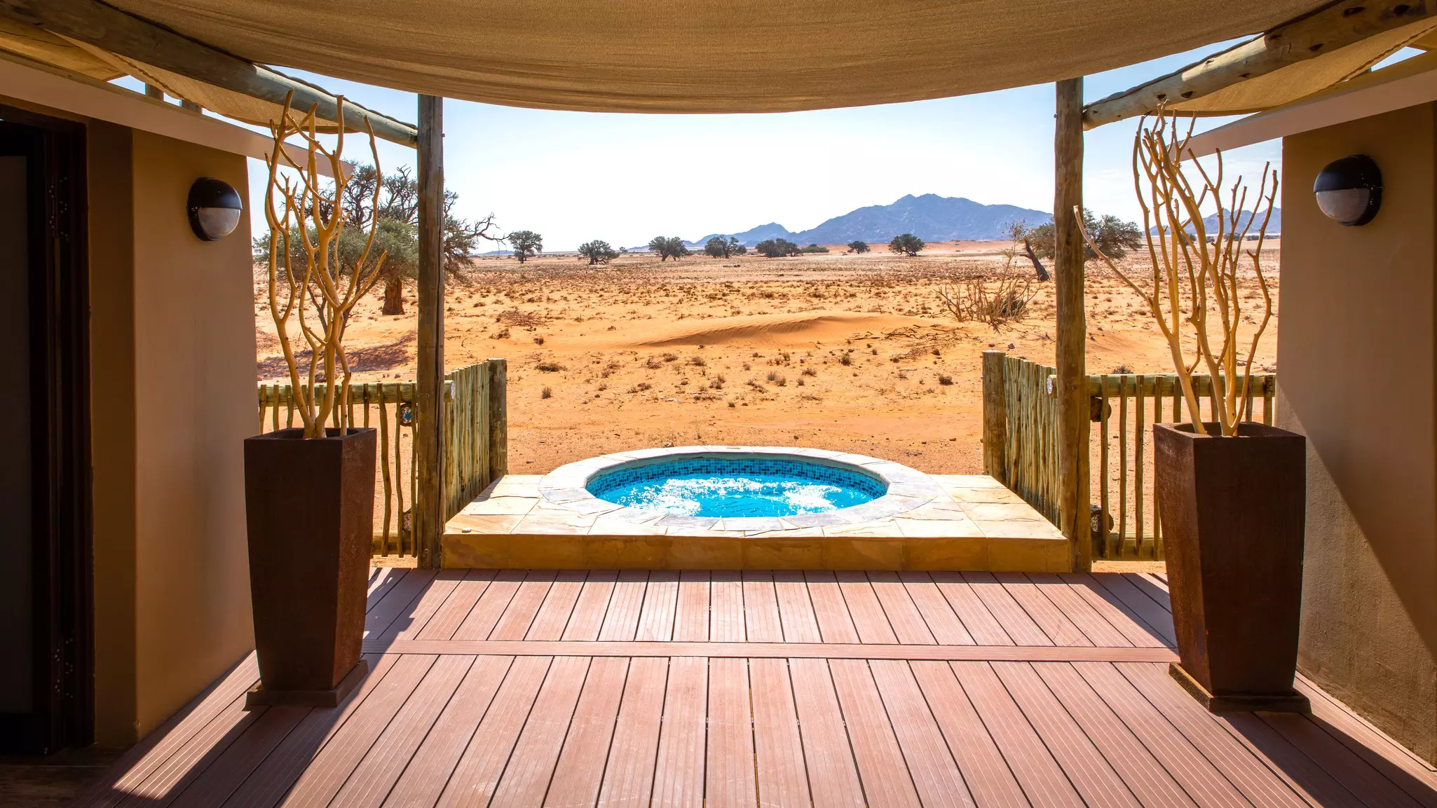 A round pool at the end of a wooden deck between two buildings is covered by an awning; a desert landscape extends beyond, with a mountain in the distance.