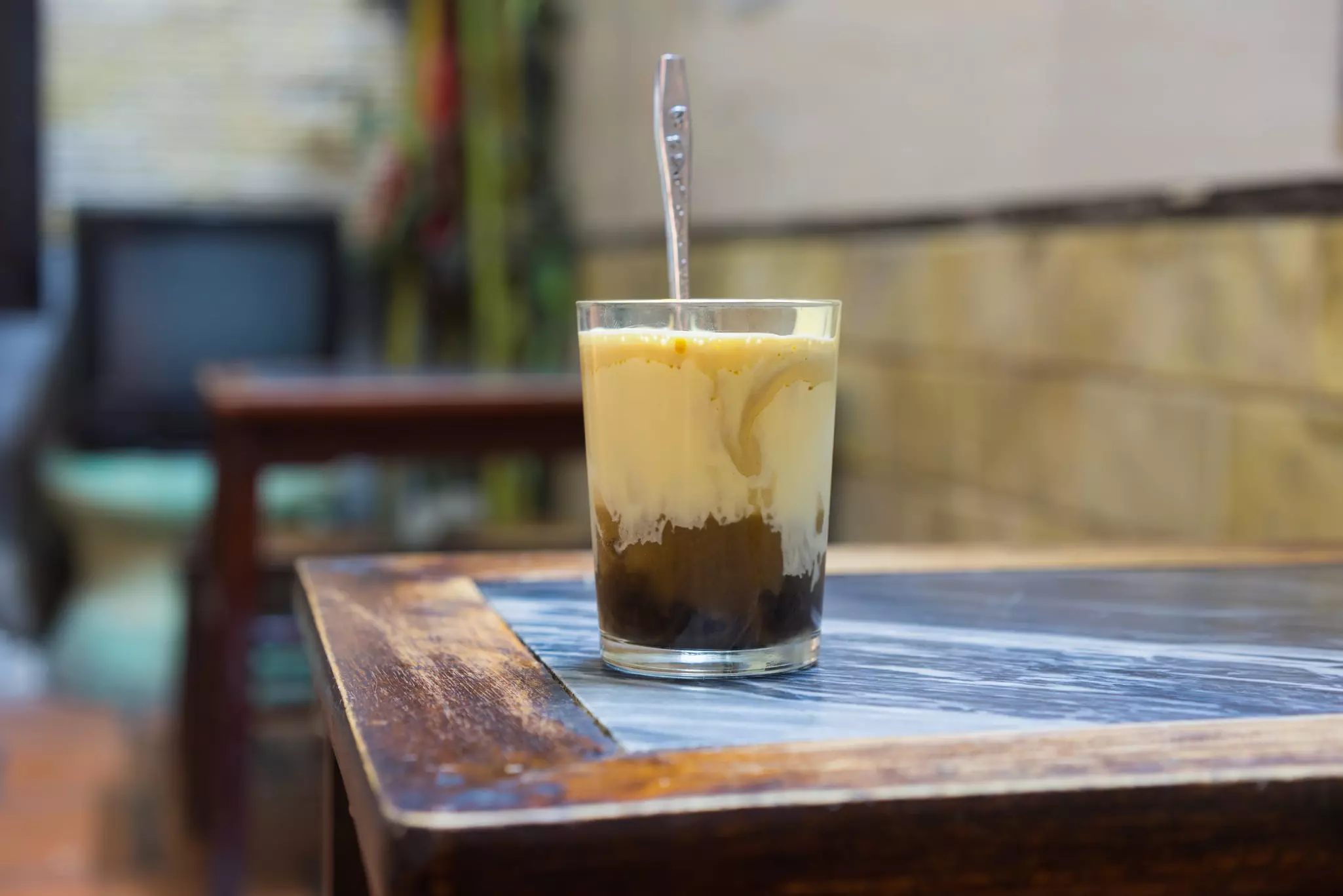 A glass with coffee and light liquid on top with a spoon sticking out on a wooden table.